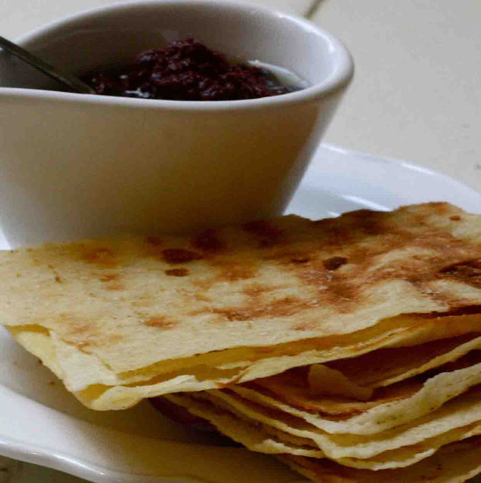 A stack of crispy flatbread is served on a white plate with a small bowl of Bella Cucina Olivada Olive Pesto beside it. A metal spoon rests in the bowl, ready to spread the bold Mediterranean flavors.