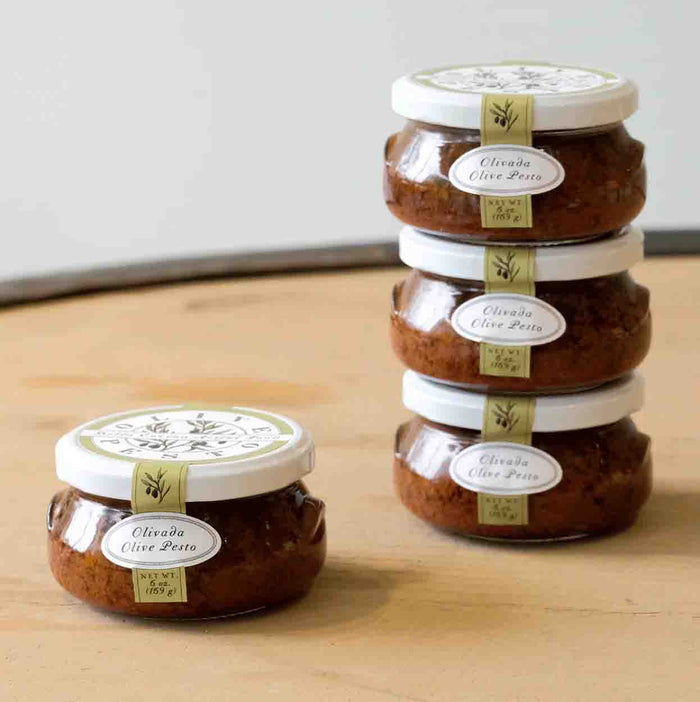 Four jars of Bella Cucina Olivada Olive Pesto, made with Kalamata olives, are arranged on a wooden surface—three stacked together and one placed separately in front.