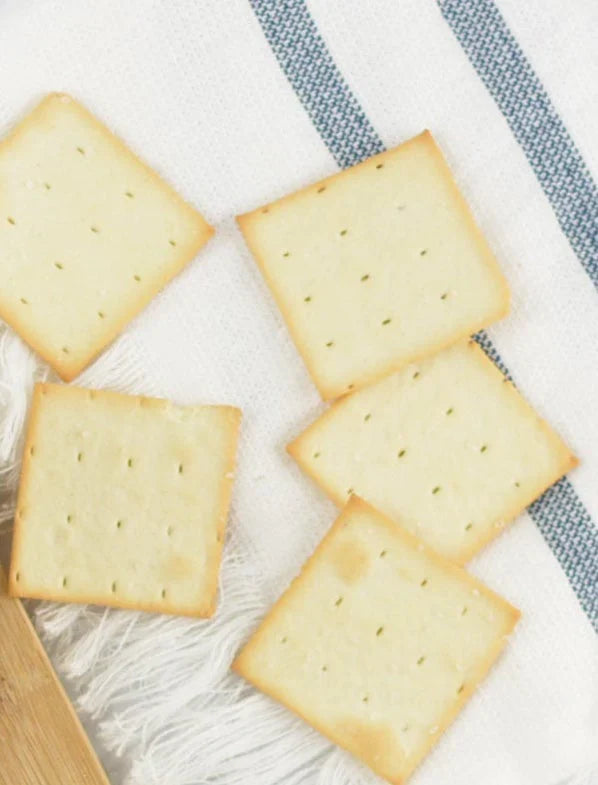 Five square Bella Cucina Olive Oil Dipping Crackers, lightly browned with small holes, are arranged on a white cloth with blue stripes near the edge of a wooden board.