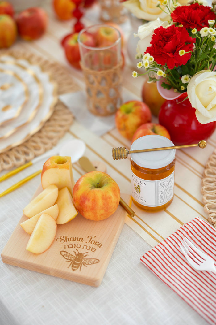 A table set with apples, Bella Cucina Organic Acacia Honey from Piedmont, Italy, flowers, and a cutting board engraved with “Shana Tova” in Hebrew to celebrate Rosh Hashanah. Sliced apple and a honey dipper rest beside a jar of this golden biodynamic honey.