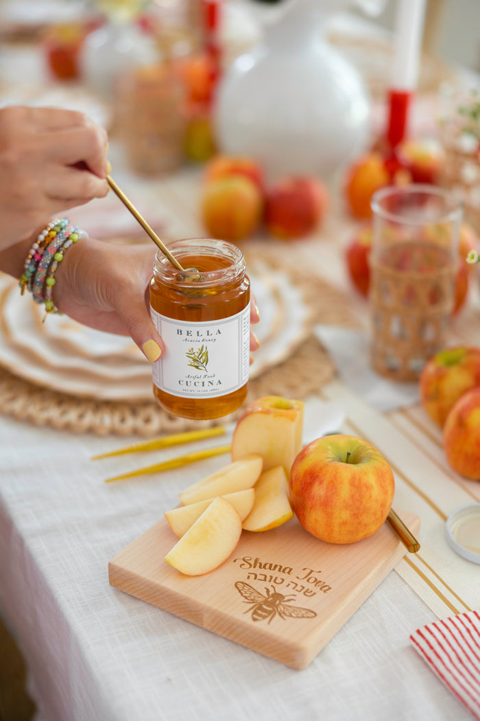 A hand dips a honey stick into a jar of Bella Cucina Organic Acacia Honey, which sits beside apples and apple slices on a wooden board engraved with Shana Tova, atop a festive table setting.