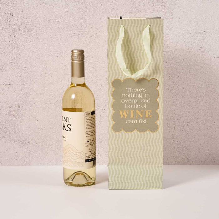 A bottle of white wine sits beside the Drinks On Me Overpriced Bottle Wine Bag, featuring wavy lines and the message: “There’s nothing an overpriced bottle of wine can’t fix!”—a fun hostess gift.