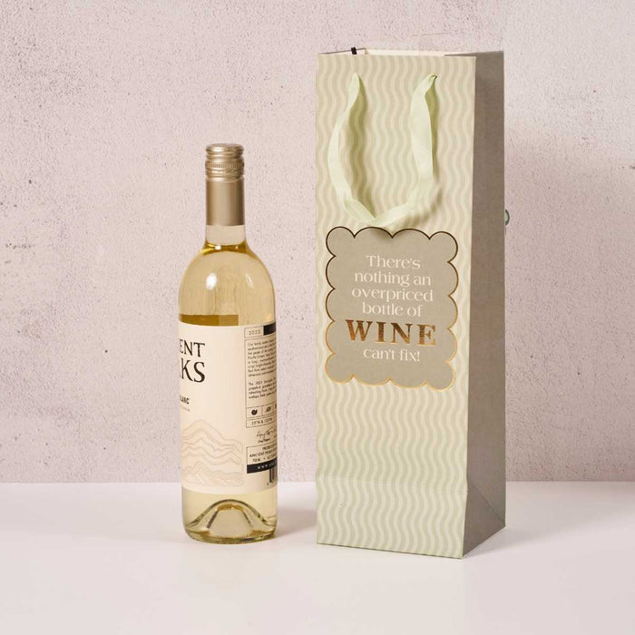 A bottle of white wine sits beside the Drinks On Me Overpriced Bottle- Wine Bag, featuring a wavy pattern and the phrase "There's nothing an overpriced bottle of WINE can't fix!"—a perfect hostess gift.