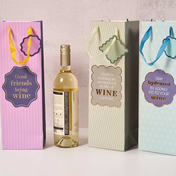 Three reusable Overpriced Bottle wine bags by Drinks On Me, in pink, light green, and blue with colorful ribbons and tags, stand by a bottle of white wine—perfect as a funny hostess gift.
