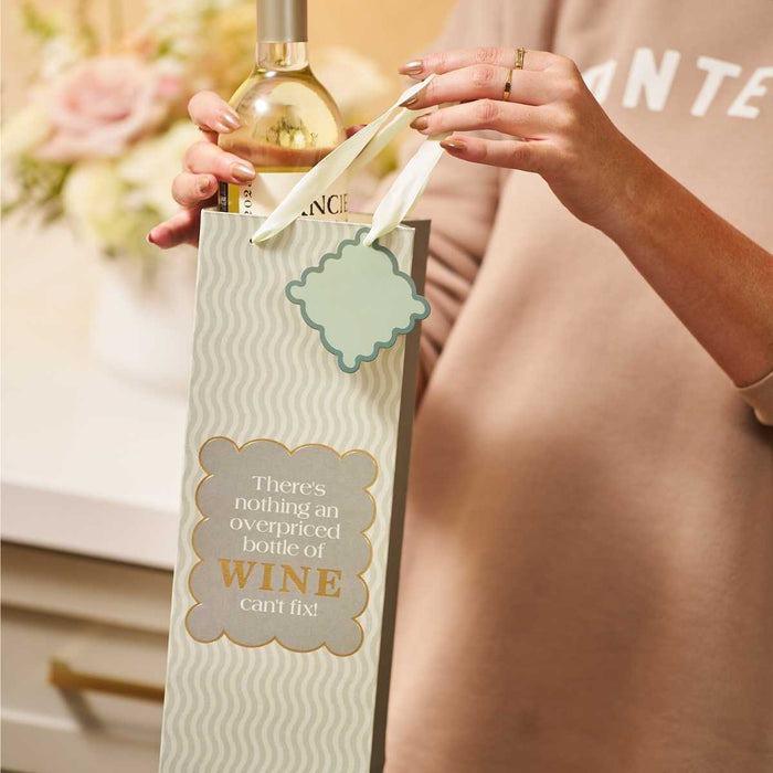 Someone holds the "Overpriced Bottle- Wine Bag" by Drinks On Me with a wine bottle inside. The bag says, "There's nothing an overpriced bottle of wine can't fix!" Perfect for hostess gifts, it's shown in a flower-filled kitchen.