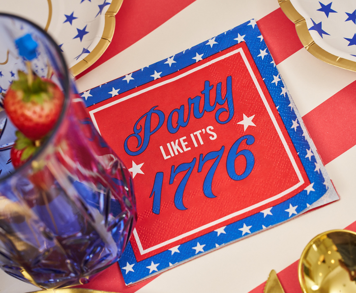 The Sophistiplate Party in the USA Cocktail Napkins (26pk) feature stars and “Party like it’s 1776”—a festive touch for patriotic tablescapes, paired with themed plates, strawberries, and gold utensils.