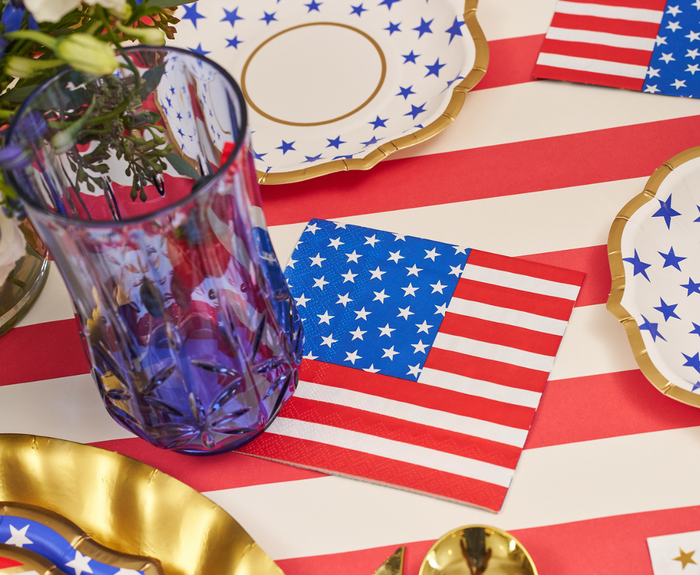 A festive table is set with gold-rimmed plates, a blue glass, and Sophistiplate Party in the USA Cocktail Napkins (26pk) featuring American flag designs on a red and white striped tablecloth.