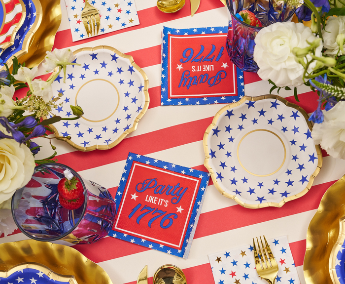 A festive table set with star plates, gold utensils, flower centerpieces, “Party Like It’s 1776” red napkins, and Sophistiplate’s Party in the USA Appetizer/Dessert Bowls on an American flag-inspired tablecloth.