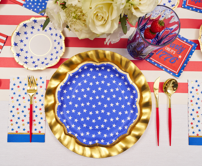Create a patriotic table with Sophistiplate’s Party in the USA Dinner Plates—gold-rimmed blue plates with white stars—paired with gold utensils, a glass of strawberries, striped red, white & blue accents, and white flowers in the background.