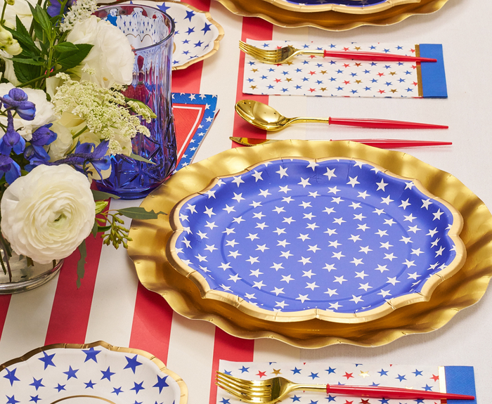 A festive table features Sophistiplate’s Party in the USA Dinner Plates, gold utensils with red handles, striped napkins, a floral centerpiece, and a red and white striped tablecloth—perfect for your patriotic Fourth of July celebration.