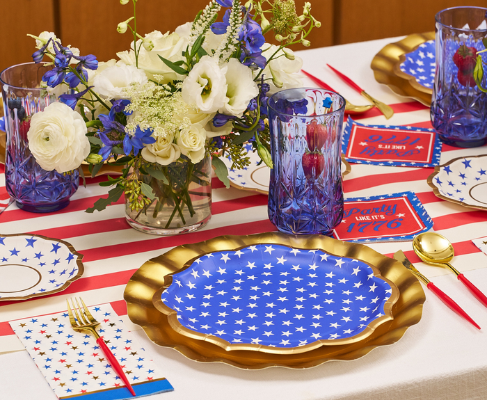 A festive table set with Sophistiplate’s Party in the USA Dinner Plates (8pk), white stars on blue, red and white striped tablecloth, blue glasses with strawberries, gold utensils, and a white and purple flower centerpiece for patriotic decor.