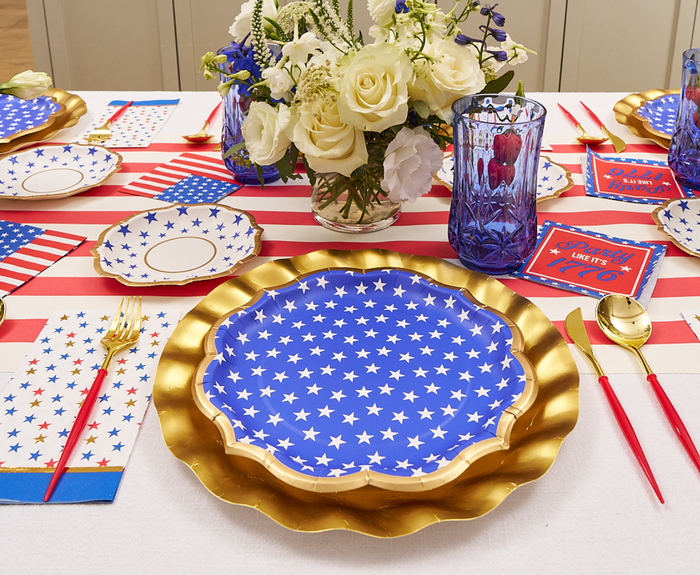 A festive table features Sophistiplate’s Party in the USA Dinner Plates, gold chargers, red-handled gold cutlery, blue glasses, white flowers as a centerpiece, and an American flag-themed runner for a patriotic July 4th look.