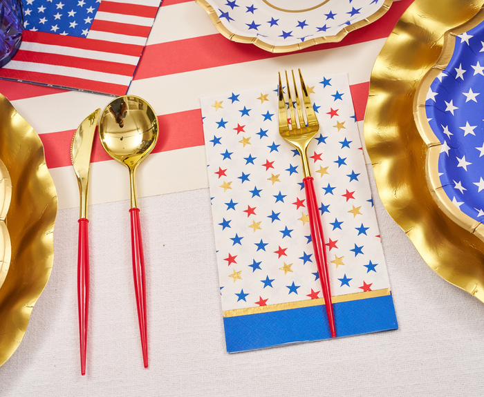 Gold-colored fork and spoon with red handles rest on Sophistiplate's Party in the USA Guest Towels, with patriotic plates and American flag decor arranged atop a white tablecloth.