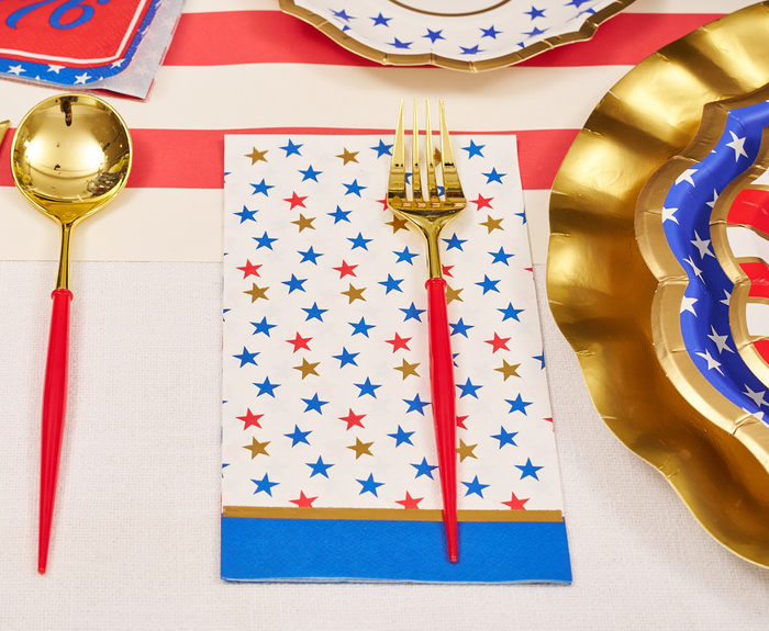A gold fork with a red handle rests on Sophistiplate’s Party in the USA Guest Towels, adorned with red, blue, and gold stars. A gold spoon and festive plates sit nearby atop a red-and-white striped tablecloth.