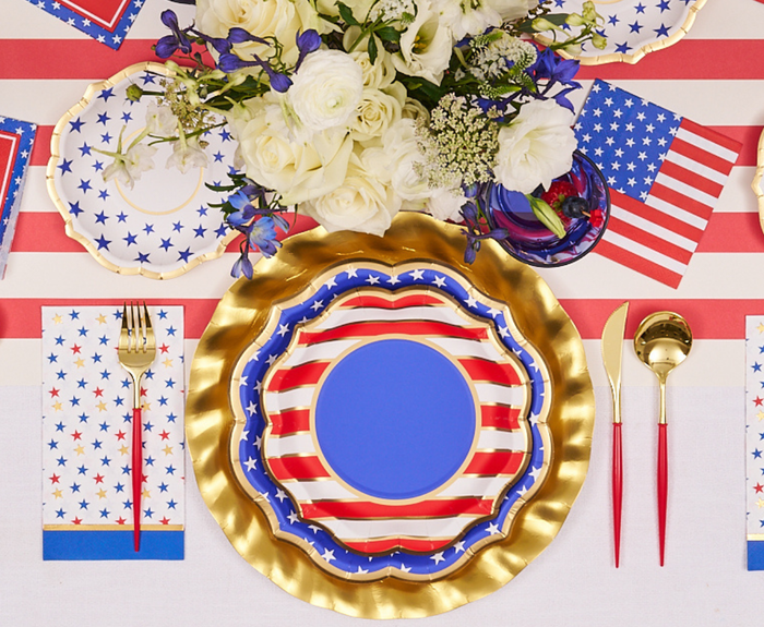 A festive table is set with Sophistiplate Party in the USA Salad Plates (8pk), featuring a red, white, and blue flag design, gold utensils, white flowers, patriotic napkins, and a striped tablecloth.