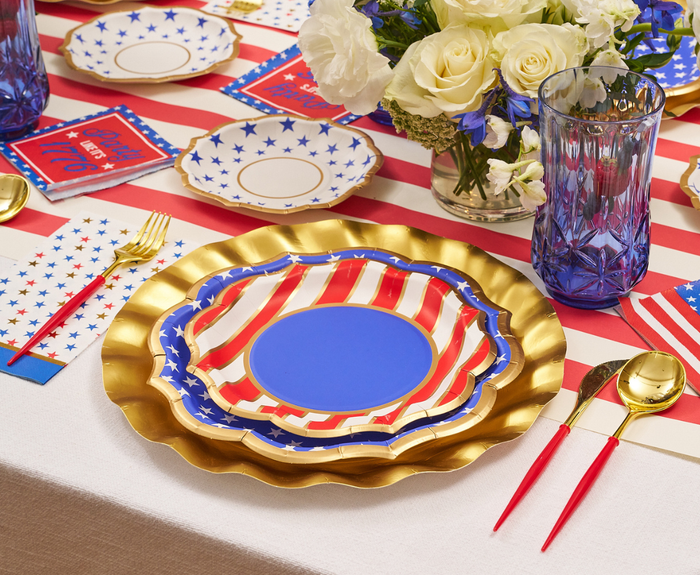 A festive table features gold cutlery, blue glasses, Sophistiplate Party in the USA Salad Plates/8pk with stars and stripes, plus matching napkins and notebooks, all accented by a bouquet of white and blue flowers.