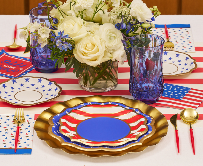 A festive table features Sophistiplate’s Party in the USA Salad Plates, gold utensils, blue glasses, white and blue flower centerpiece, plus American flag napkins and placemats.