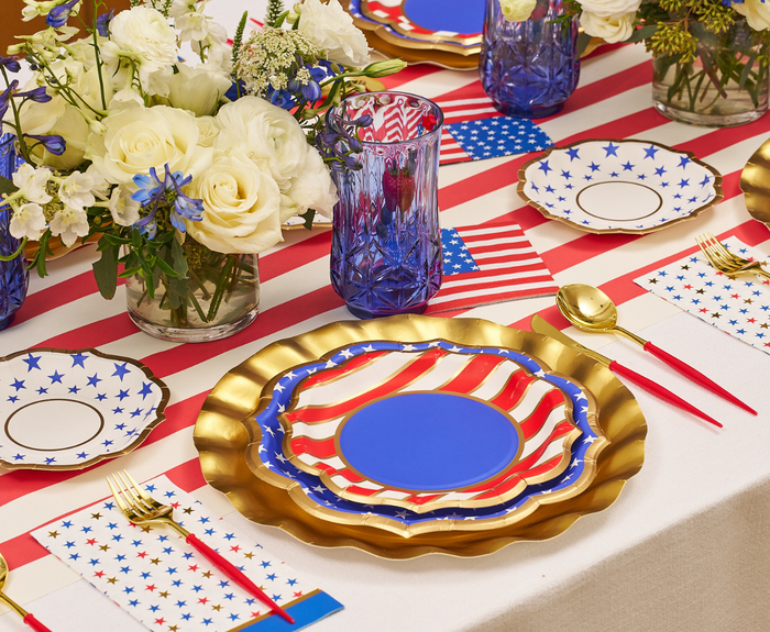 A festive table features Sophistiplate’s Party in the USA Salad Plates (8pk), gold-rimmed blue glasses, gold utensils with red handles, and floral arrangements atop a red-and-white striped runner.