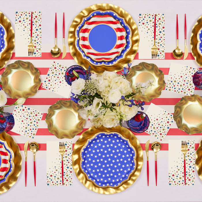The Party in the USA Table Setting by Sophistiplate LLC includes gold-rimmed American flag plates, gold and red cutlery, patriotic napkins, and a white flower centerpiece—ideal for Fourth of July or as a party in a box.