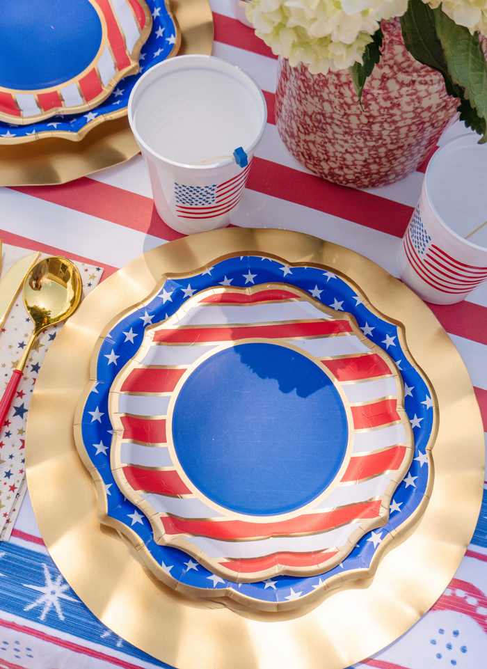 The Party in the USA Table Setting by Sophistiplate LLC features patriotic plates and cups, gold cutlery, and a striped tablecloth with white flowers—perfect for a Fourth of July celebration or an all-in-one party box.