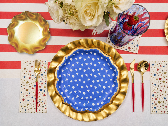 The Party in the USA Table Setting by Sophistiplate LLC features patriotic plates with white stars and a gold rim, on a red and white striped tablecloth, paired with gold utensils, star napkins, and a floral centerpiece.