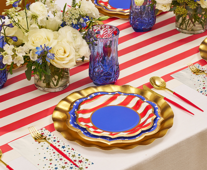 The Party in the USA Table Setting by Sophistiplate LLC includes patriotic plates, gold chargers, colorful-handled gold flatware, star napkins, blue glasses, and white flowers—everything you need for a stylish celebration.