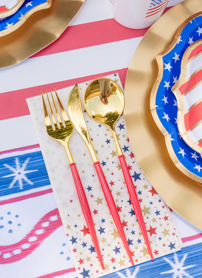 Sophistiplate LLC’s 4th of July Table for 16+: Party in the USA set features gold flatware with red handles on a star-patterned napkin and patriotic plates, arranged with festive decorations in red, white, and blue.