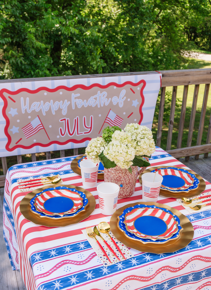 A 4th of July table set for 16+ features Sophistiplate LLC’s “Party in the USA” party pack with disposable plates, American flag cups, gold cutlery, a floral centerpiece, and a “Happy Fourth of July” banner.