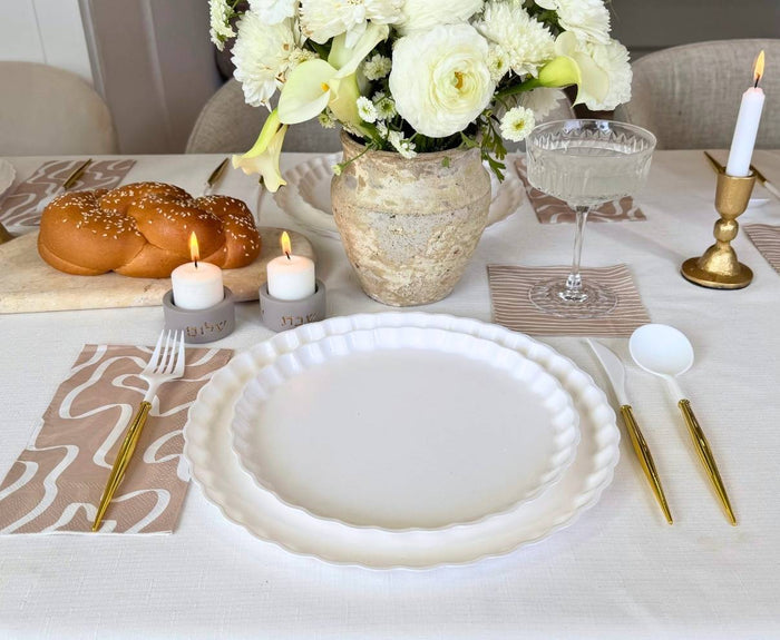 A meal table is set with Sophistiplate’s Eco-Friendly Table Setting-White, including disposable plates, gold cutlery, a wine glass, patterned napkins, lit candles, white flowers in a vase, and challah bread on a cutting board.