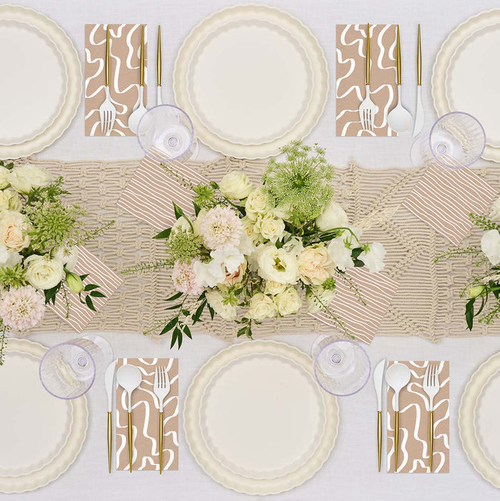 The Sophistiplate Eco-Friendly Table Setting-White seats six and features disposable plates, gold cutlery with patterned napkins, clear wine glasses, and floral centerpieces in white and pale pink on a beige woven runner.