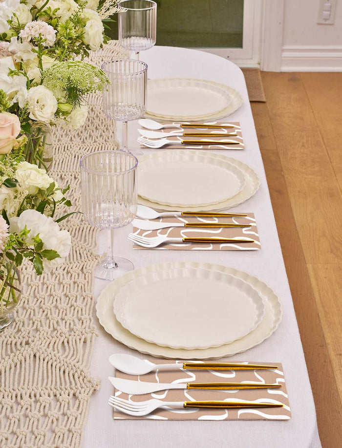 A Sophistiplate Eco-Friendly Table Setting- White features disposable plates, gold and white cutlery, patterned napkins, clear ribbed glasses, and a woven runner adorned with white and cream florals.