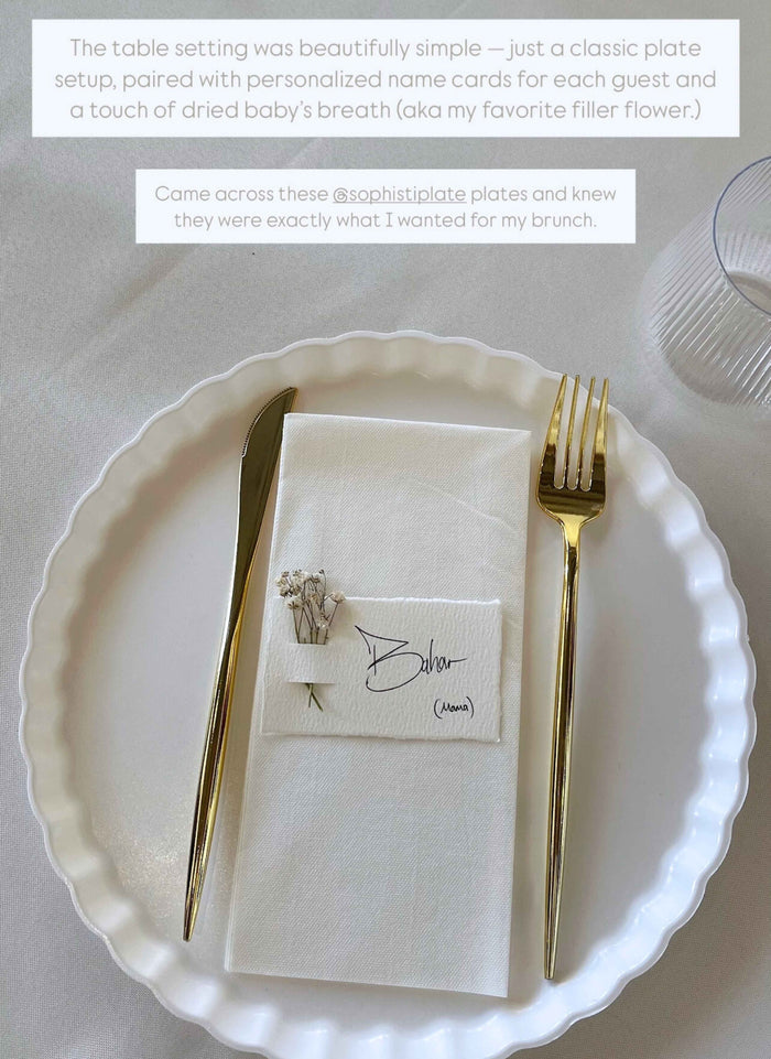 The Sophistiplate Eco-Friendly Table Setting-White features a white scalloped plate, gold knife and fork, napkin, and place card with dried baby's breath beside a clear ribbed glass on a light tablecloth.