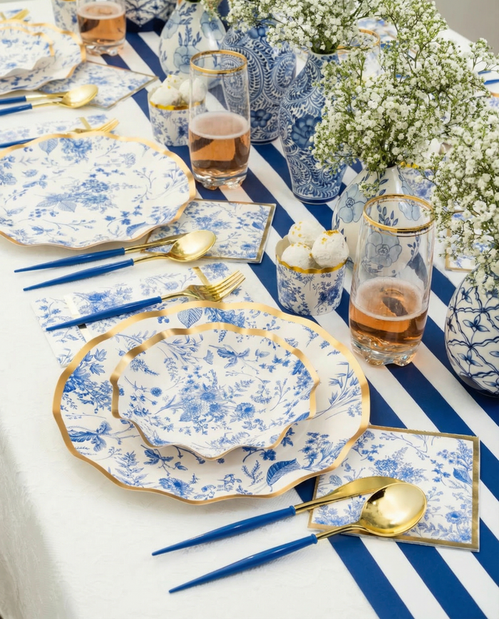 The Passover Tablescape for 16: Timeless by Sophistiplate LLC features blue and white floral paper plates, gold and blue cutlery, pink drinks in glasses, and vases of baby’s breath on a white tablecloth with a blue striped runner.