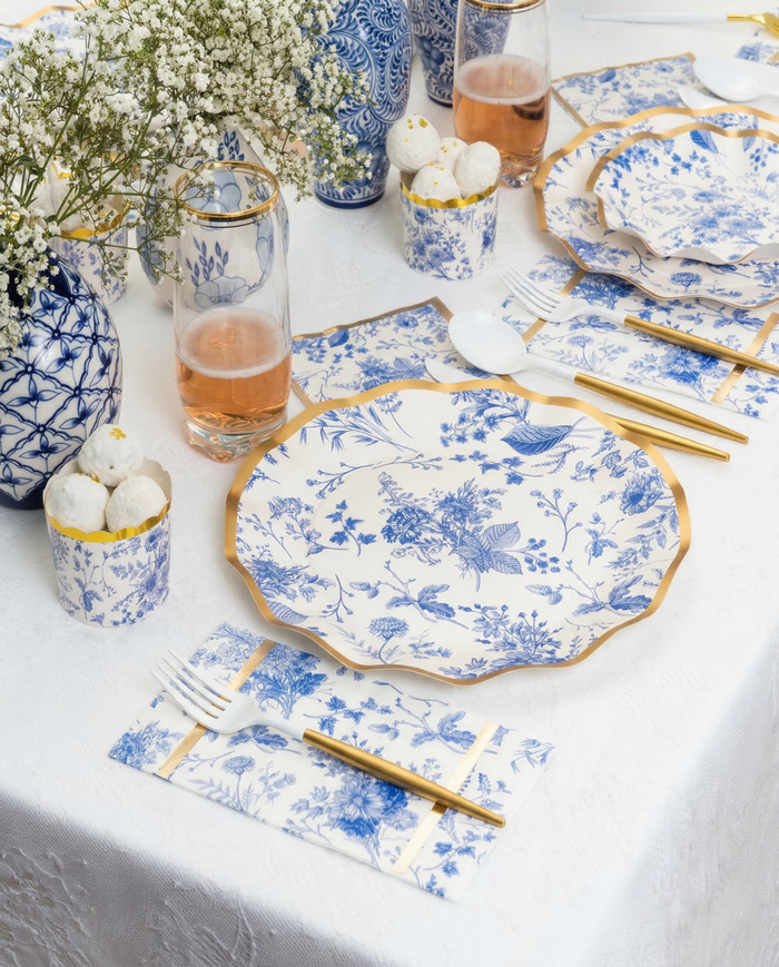 Create an elegant Passover tablescape for 16 with Sophistiplate LLC’s Timeless set, featuring blue and white floral paper plates, gold-rimmed cutlery, pink drinks in glasses, white treats in cups, and baby's breath vases on a crisp white tablecloth.