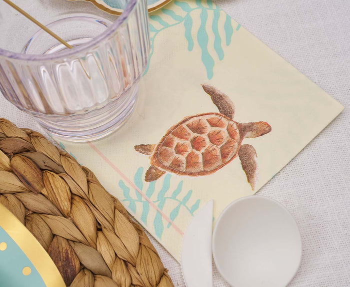 A clear glass rests on a Sophistiplate Pastel Sea Cocktail Napkin, adorned with a sea turtle design. Nearby are a woven placemat, white plastic spoon, and gold-rimmed plate on a pale tablecloth. Perfect for coastal-themed parties.
