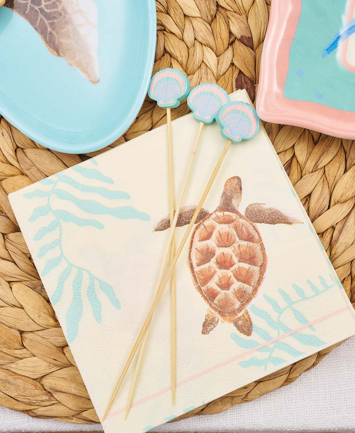 Sophistiplate’s Pastel Sea Cocktail Napkins (20pk) are displayed on a woven mat with three blue seashell cocktail picks, alongside light blue and pink plates for a charming coastal-themed table setting.