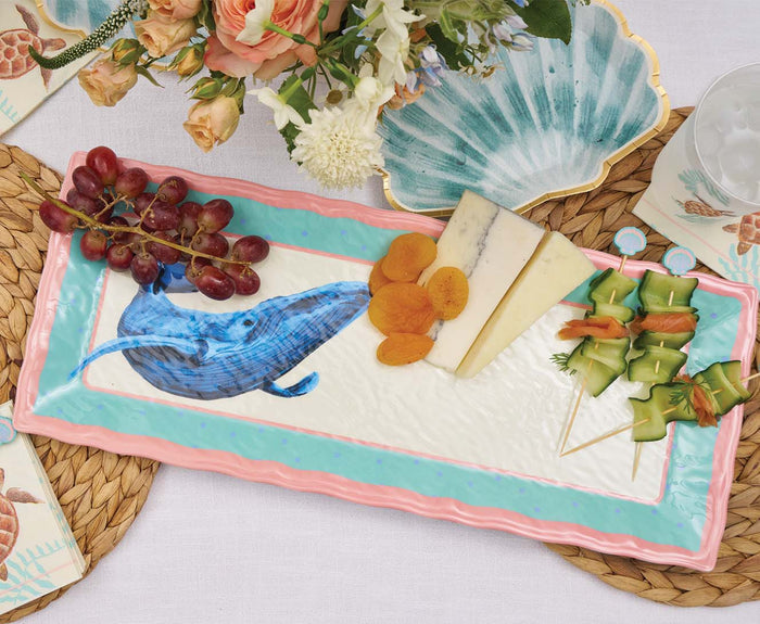 A Sophistiplate Pastel Sea Die Cut Shell Salad Plate holds grapes, dried apricots, cheese slices, and veggie skewers. Nearby are floral arrangements and a glass of ice water on a woven placemat.
