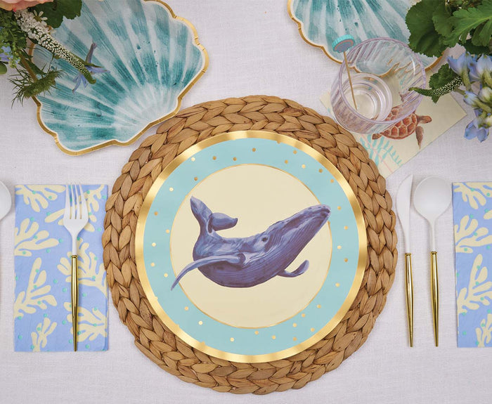 A table setting with a woven placemat, a whale plate, gold and white cutlery, a glass, Sophistiplate Pastel Sea Die Cut Shell Salad Plates (8pk), and blue coral-patterned napkins on a white tablecloth.