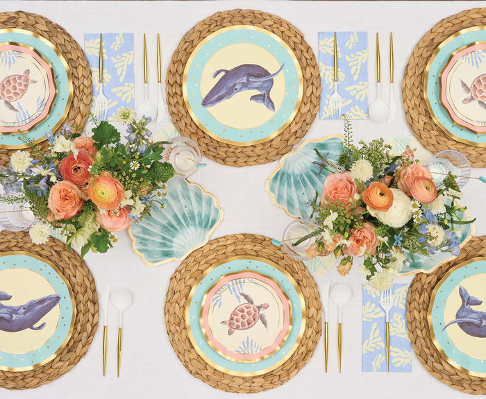 A table set for six with woven placemats, coastal party plates with dolphins and turtles, gold flatware, blue coral napkins, Sophistiplate Pastel Sea Die Cut Shell Salad Plates, and floral centerpieces in peach, white, and orange.