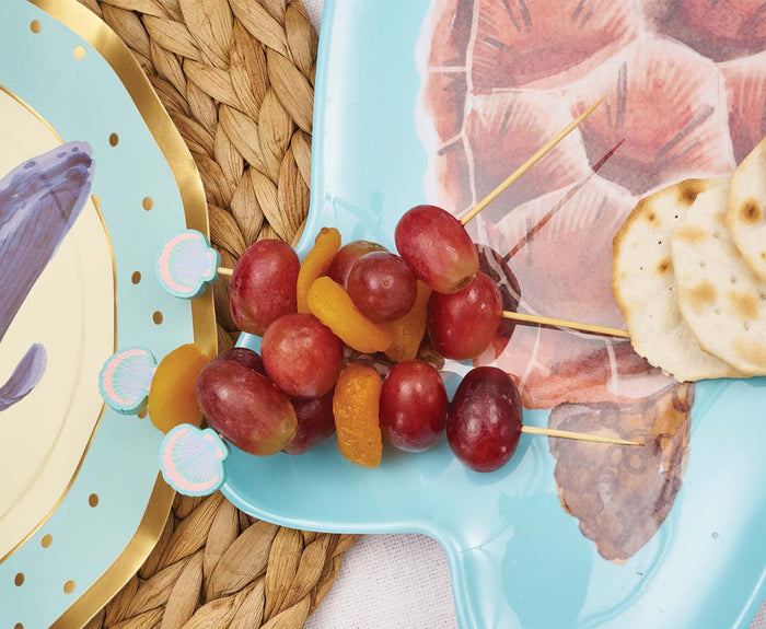 Simply Serv'd Pastel Sea Large Cocktail Picks (30pk) skewer red grapes and dried apricots, arranged on a blue plate with a sea turtle design, served beside round crackers and a woven placemat.