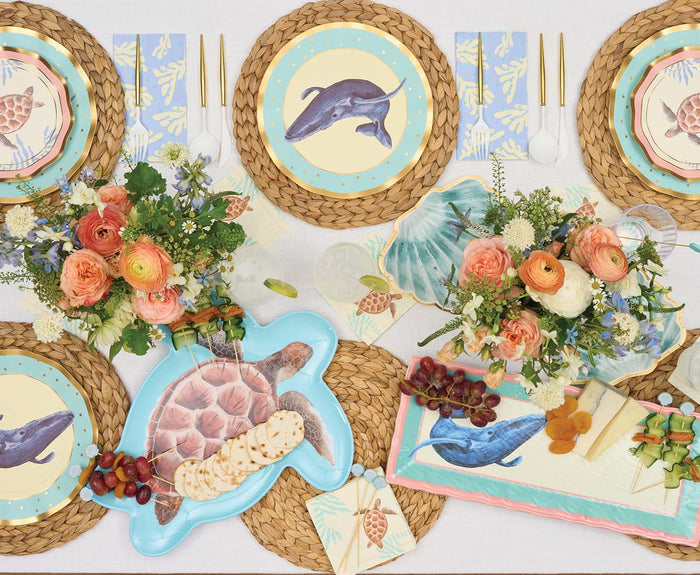 A table set with wicker placemats, whale and turtle sea-themed plates, a Sophistiplate Pastel Sea Melamine Serving Tray, floral centerpieces, gold utensils, and platters of cheese, crackers, grapes, and snacks in a light festive style.