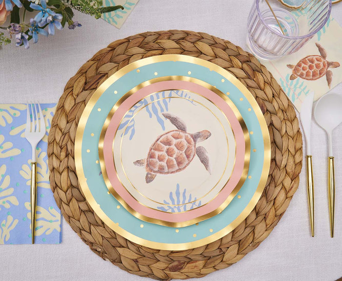 A place setting with a woven placemat, three Sophistiplate Pastel Sea Salad Plates (featuring ocean-themed designs and a sea turtle), gold utensils, a cup with purple stripes, and coral-patterned napkins. Flowers appear in the upper left corner.