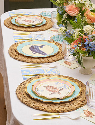 A table set with woven placemats, Sophistiplate Pastel Sea Salad Plates featuring whale and sea turtle designs, gold utensils, clear glasses, and a floral centerpiece with white and orange flowers.