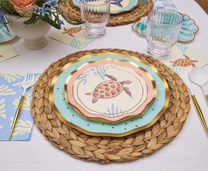 A table setting featuring Sophistiplate’s Pastel Sea Salad Plates with a sea turtle design (8-pack), paired with a woven placemat, gold cutlery, clear glassware, floral napkins, and a flower arrangement on a white tablecloth.