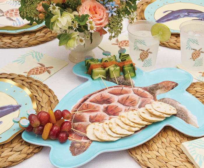 The Sophistiplate Pastel Sea Turtle Melamine Serving Platter displays fruit skewers, grapes, and crackers on a woven placemat alongside plates, napkins, turtle-themed cups, and a floral centerpiece—ideal ocean-inspired serveware.