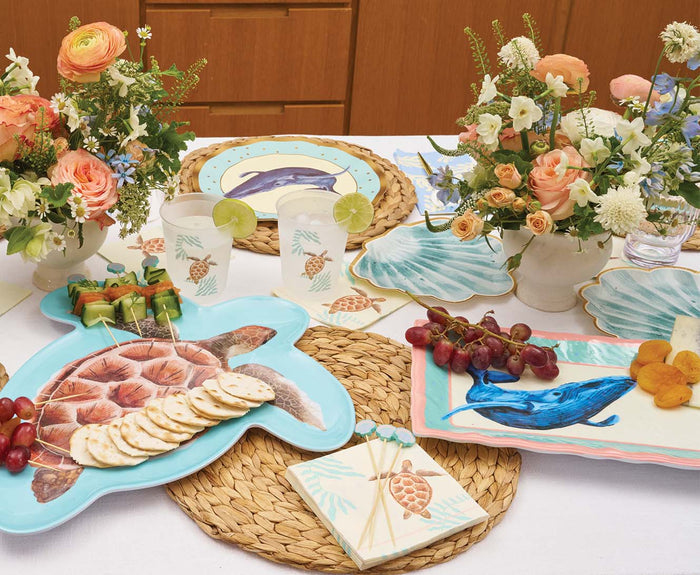 A table set with sea animal-themed plates, floral centerpieces, snacks, and drinks is brightened by the Sophistiplate Pastel Sea Turtle Melamine Serving Platter, adding a cheerful, beachy touch.