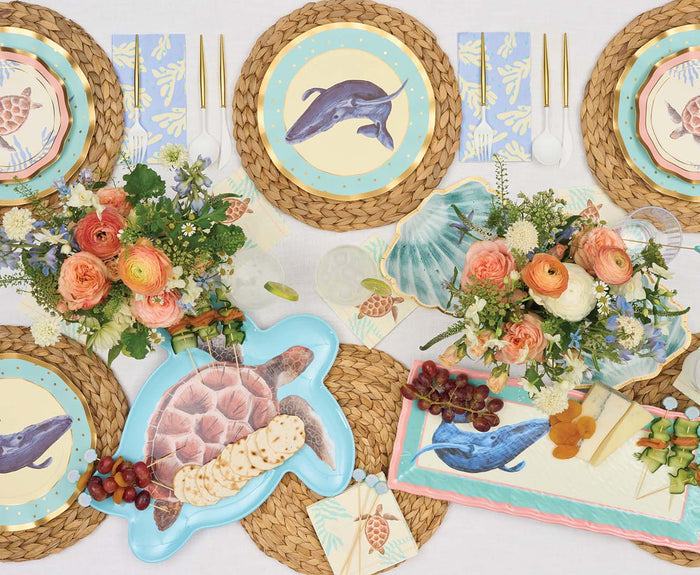 A tabletop set features woven placemats, plates with whale and turtle designs, floral centerpieces, gold utensils, leafy napkins, and Sophistiplate's Pastel Sea Turtle Melamine Serving Platter with grapes, crackers, and cheese.
