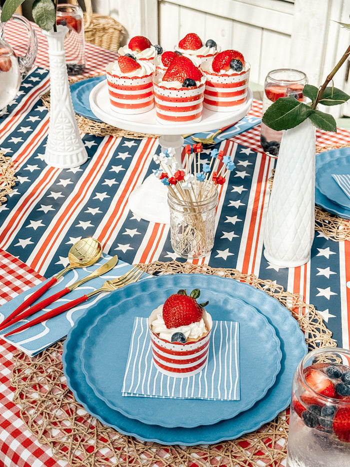 A festive table features Sophistiplate LLC's Patriotic French Blue plates, gold and red cutlery, red-white-blue décor, berry-topped cupcakes on striped napkins, drinks, a star-patterned tablecloth, and eco-friendly disposable plates.