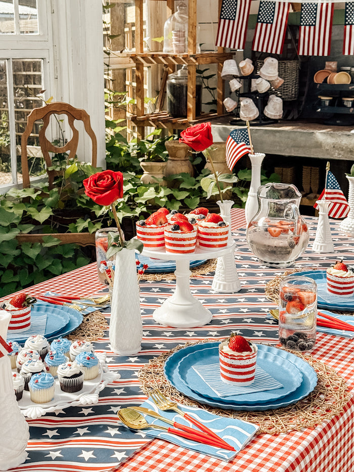 Create a festive celebration with the Patriotic French Blue collection from Sophistiplate LLC—featuring red, white, and blue desserts, American flags, cupcakes, compostable bamboo plates, vases with red roses, and a starry checked tablecloth.