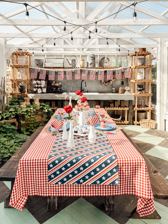 A festive outdoor dining table showcases Sophistiplate LLC's Patriotic French Blue collection, with stars-and-stripes runners, red flowers, American flags, eco-friendly plates, all beneath string lights in a white pergola.